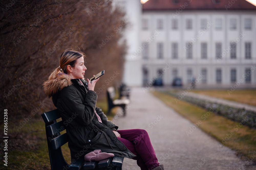 Fototapeta premium Woman converses on a smartphone while seated on a park bench, a wisp of breath visible in the cool air