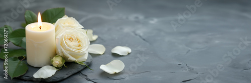 White candle with a rose and flowers petals on grey stone panoramic background with copy space, funeral web banner photo