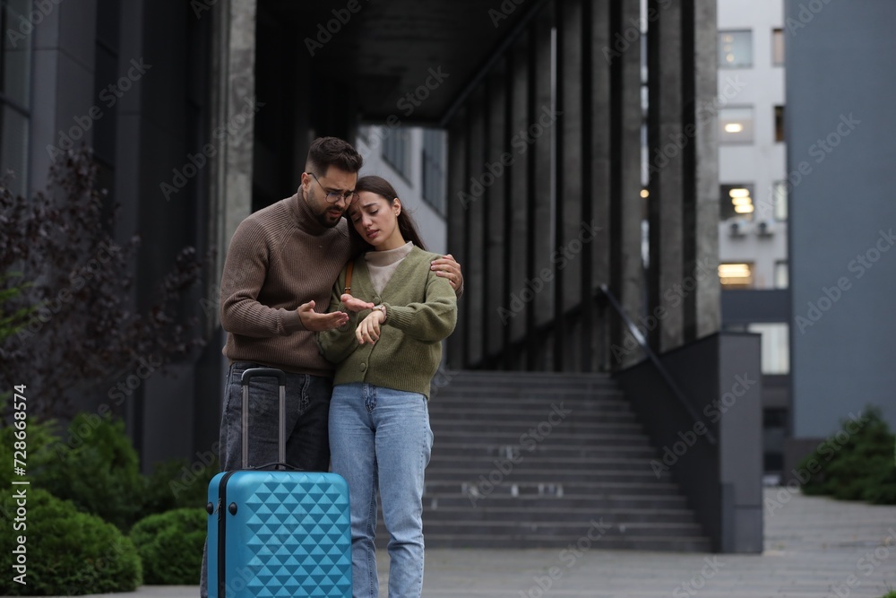 Being late. Worried couple with suitcase looking at watch near building outdoors, space for text Being late. Worried couple with suitcase looking at watch near building outdoors, space for text