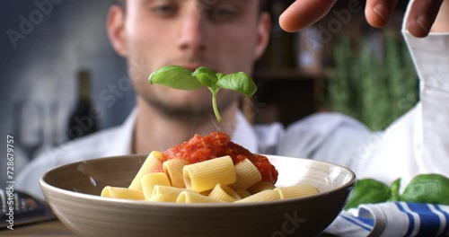Super slow motion of a professional chef is putting on  fresh green basil leaf on traditional Italian dish of pasta penne with red tomato sauce in restaurant kitchen at 1000 fps. photo