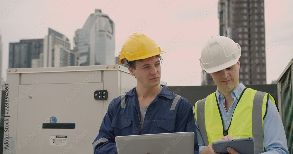 Two male engineers in hard hats reviewing data on a laptop on a construction site, with skyscrapers in the background Two male engineers in hard hats reviewing data on a laptop on a construction site, with skyscrapers in the background
