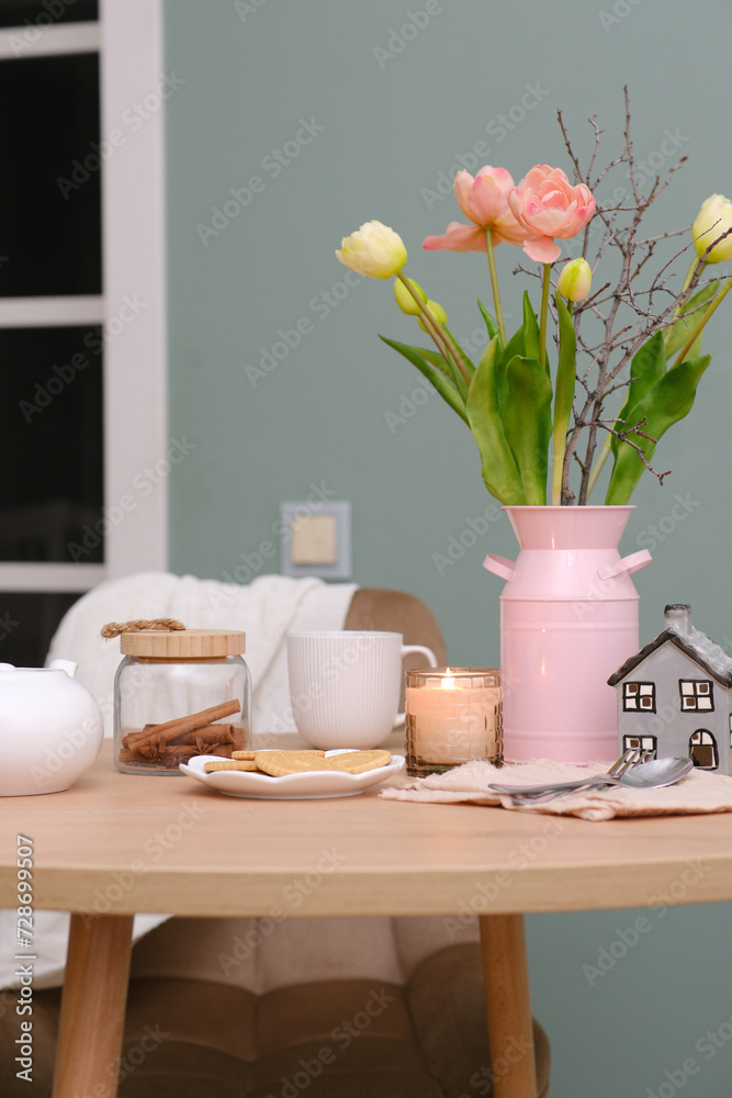 Home spring interior in living room. Pink vase with tulips and branches,hot teapot, burning candle, cup of tea on round wooden table in cosy living room.
