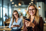 A cheerful and smiling young successful female businesswoman standing with colleague looking at smartphone in modern office and coworking space.