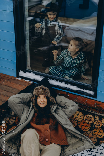 young woman dressed in warm country clothes on the background of a country house for vacation with two chilren enjoying the winter view of the village photo