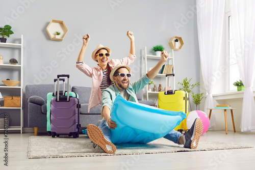 Funny excited happy couple in hats and sunglasses having fun sitting on inflatable mattress ready for holiday trip in living room at home with packed luggage. Vacation and summer journey concept.