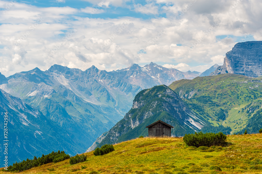 A small wooden hut perched on a green hill high in the mountains, picturesque alpine landscape in the background. Scenic hike on Blaser mountain in summer, Tyrol, Austrian Alps.