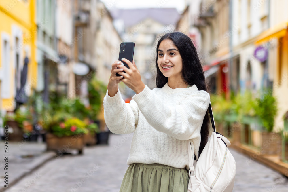 A walk through the city and a trip of an Indian young woman who stands on the street with a backpack and takes a selfie on the phone, talks with a smile on a video call, takes photos of landmarks