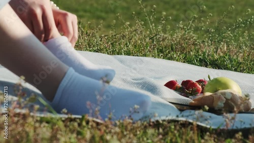 Woman in jeans puts on sock sitting on blanket during picnic. Summer picnic with juicy fruits and fresh pastry on sunny day photo
