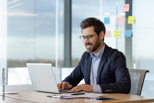 Joyful successful businessman at work inside office, man in business suit typing on keyboard, senior experienced professional investor near window smiling, satisfied with achievement results.
