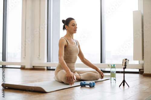 Peaceful senior caucasian woman performing padmasana in lotus pose engaging in healthy lifestyle. Famous blogger shooting video content on smartphone for own followers about meditation at studio.