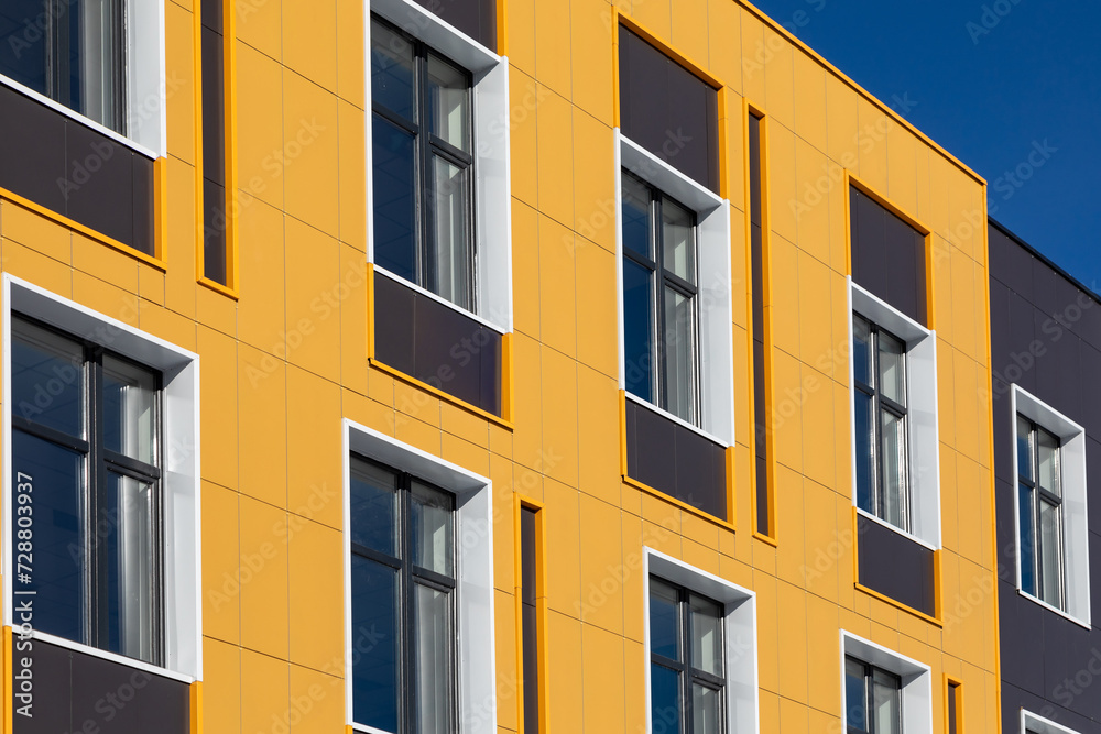 Part of building with metal facades of different colors and windows of different shapes against blue sky. Commercial real estate, office building, school