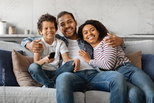 Cheerful young black family sitting on sofa, holding remote control and smiling