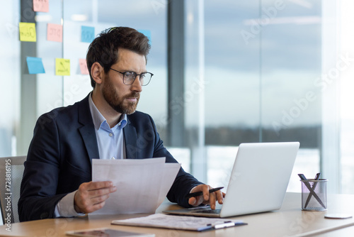 Serious and focused financier accountant on paper work inside office, mature man using calculator and laptop for calculating reports and summarizing accounts, businessman at work in suit.