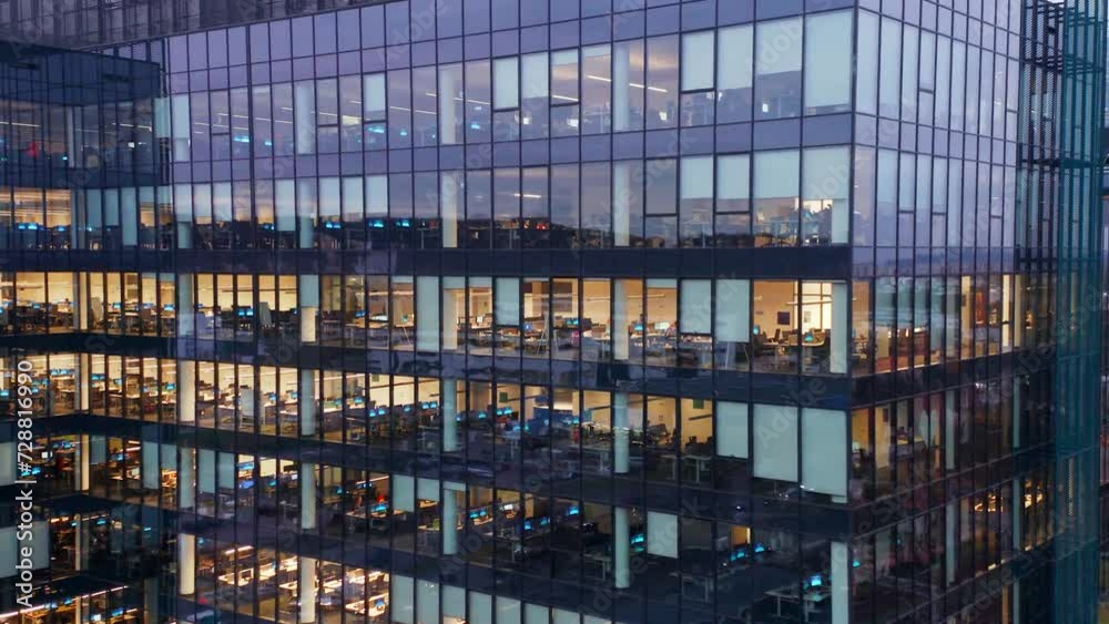 Cinematic drone shot office windows in a glass skyscraper at night close-up. People working in office building in the night, building lights. Workers at office building at night, view trough a window