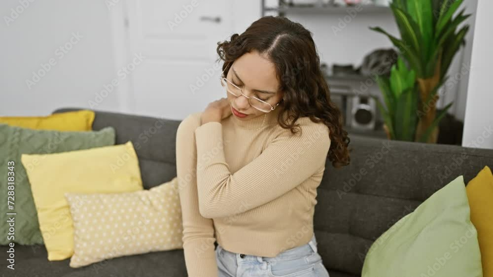 Woman holding shoulder in pain sitting on a couch indoors with decorative pillows around. Woman holding shoulder in pain sitting on a couch indoors with decorative pillows around.