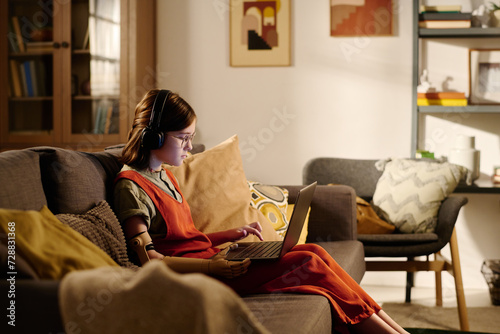 Cute pre-teen girl with disability looking at laptop screen and listening to music in headphones or watching online video while sitting on couch