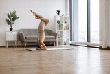 Brunette staying in headstand pose at bright commodious living room. Fit and slender lady doing yoga exercises in beige tight leggings and top in front of french windows.