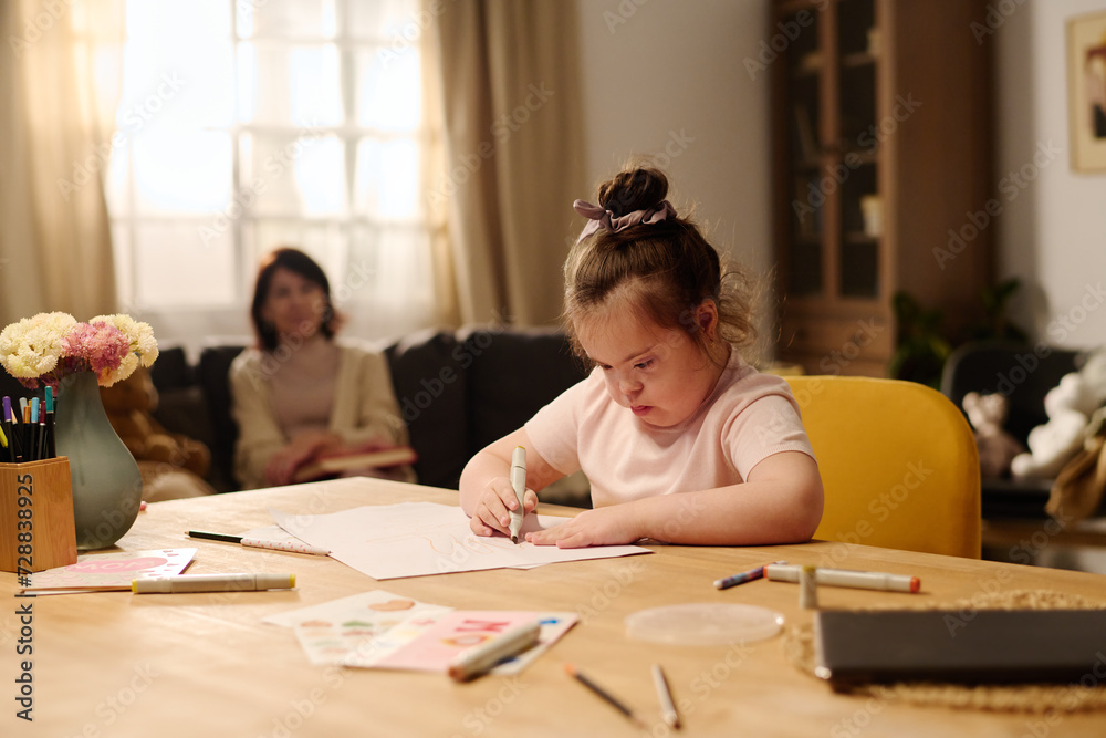 Adorable girl with mental disability sitting by table and drawing with highlighters at leisure against her mother sitting on couch Adorable girl with mental disability sitting by table and drawing with highlighters at leisure against her mother sitting on couch