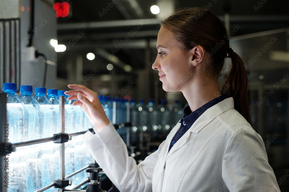 Young female quality inspector in labcoat standing in front of assembly line with capped bottled drinks and examining plastic packaging Young female quality inspector in labcoat standing in front of assembly line with capped bottled drinks and examining plastic packaging