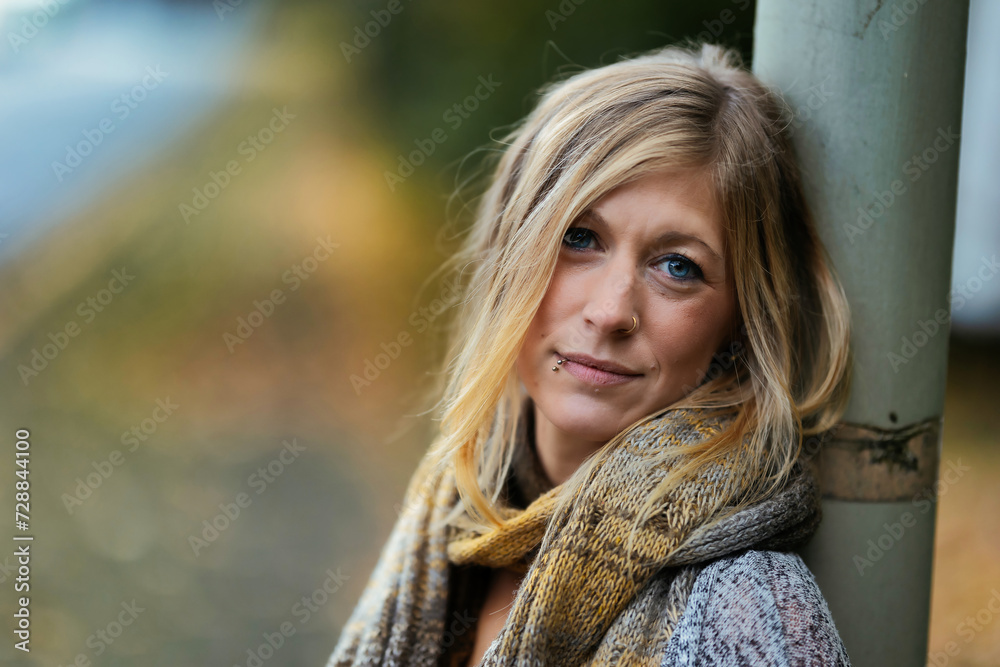 Naklejka premium Young blonde woman in a knitted look with piercing and knitted scarf leaning against a lamppost.