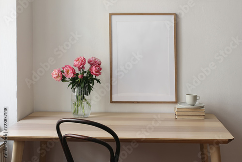 Elegant interior still life. Blank vertical picture frame mockup. Vase with pink peonies flowers. Cup of tea, coffee on books. Wooden table, desk. Romantic home breakfast. White wall background.