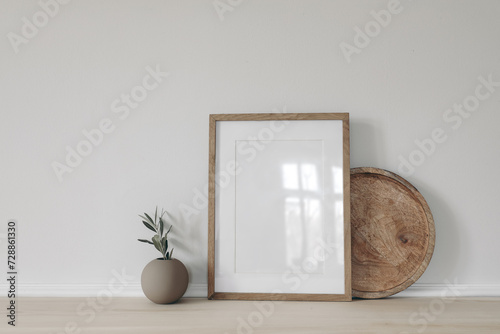 Blank vertical picture frame mockup, poster display. Ball vase with olive tree branch. Round wooden tray on table, desk. Minimal rustic home. Scandinavian interior. White wall background. photo