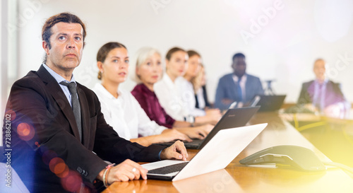 Serious middle-aged white brunette businessman sitting among colleagues at conference table in meeting room, intently listening to presentation during corporate briefing