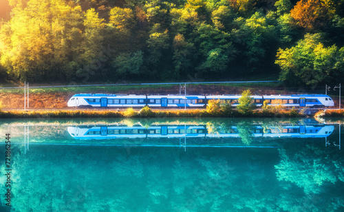 Beautiful blue modern high speed train and river in alpine mountains at sunrise in spring. Passenger train, reflection in water, railroad, lake, green forest in summer. Railway station in Slovenia