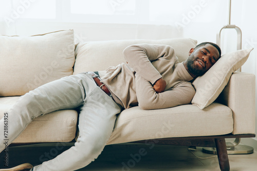 Man resting on a comfortable modern sofa in his living room, smiling as he enjoys a relaxed weekend at home The African American guy is alone, holding a smartphone and thinking, with a background of a