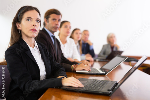 Portrait of absorbed white dark-haired female office worker sitting with colleagues in conference room and interestedly watching speaker's new project presentation during corporate group meeting