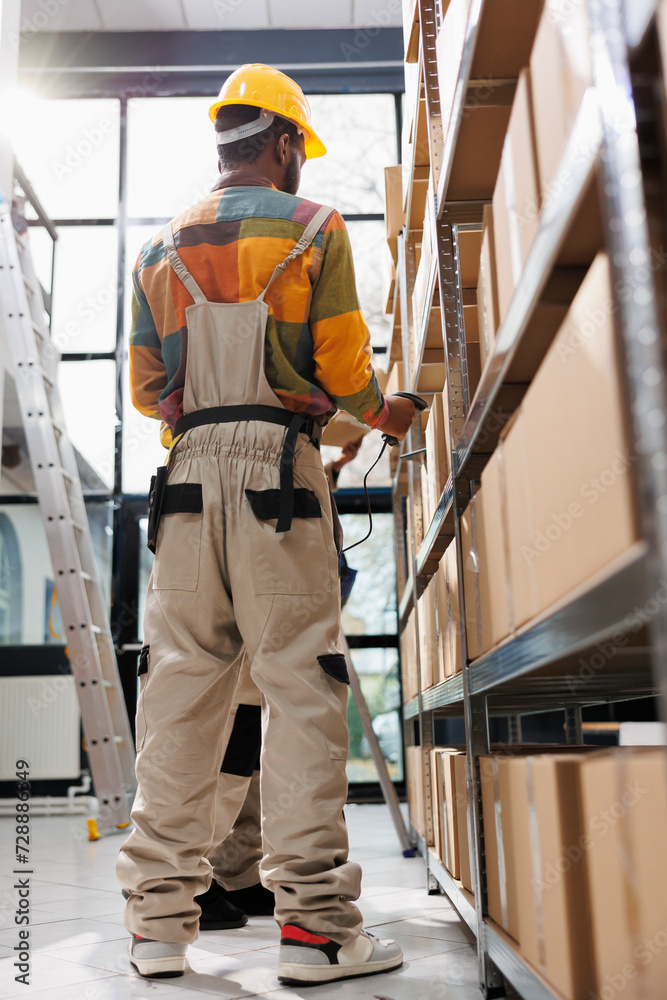 Warehouse employees managing products storing using barcode scanner. African american industrial storehouse employees wearing safety workwear standing near cardboard boxes shelf and scanning parcel Warehouse employees managing products storing using barcode scanner. African american industrial storehouse employees wearing safety workwear standing near cardboard boxes shelf and scanning parcel