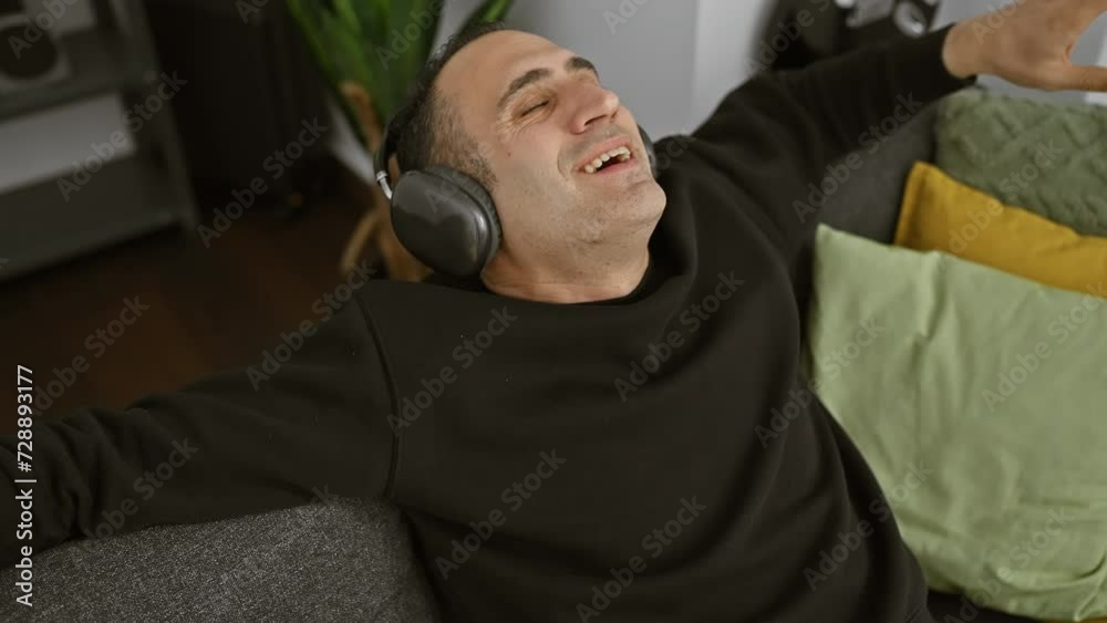 A relaxed man at home enjoying music with closed eyes and headphones, surrounded by couch pillows. A relaxed man at home enjoying music with closed eyes and headphones, surrounded by couch pillows.
