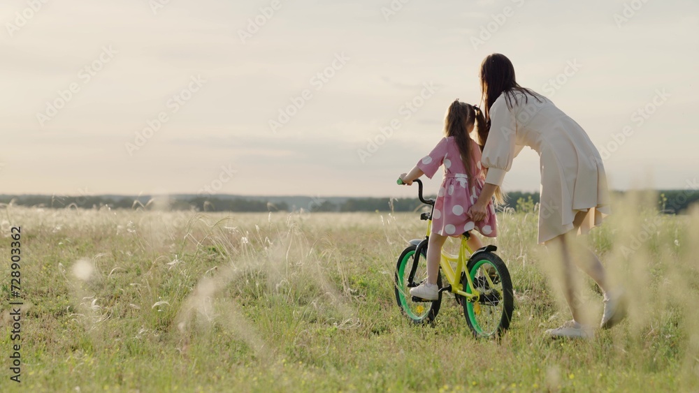 Elder sister teaches his little sister to ride bike in field, sunset. Mom teaches little girl to ride children bike in park spring. Happy family. Mommy kid play together outdoors. Pedaling bike. Child
