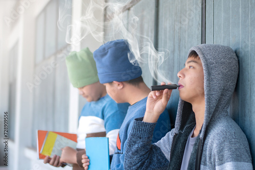 Asian preteen sitting in front of windows of school building and smoking e-cigarette not far from his friends, bad habit and behavior of young people in drug concept, soft focus, white smoke edited.