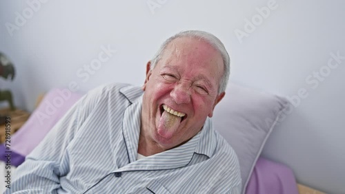 Cheerful senior man in pyjamas sitting on bed in bedroom, sticking tongue out in a hilarious expression of unadulterated joy. emotionally charged portrait in indoor setting.