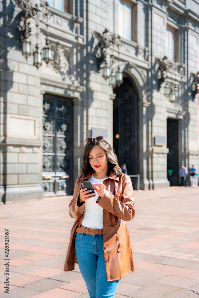 Fototapeta premium Happy young mexican woman in embroidered top looking at screen of smartphone while standing in front of blurred Munal museum in downtown Mexico City and taking selfie in daylight