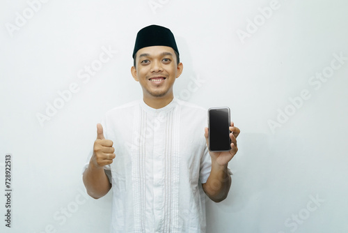 Indonesian Asian muslim man smiling and showing smartphone with thumb up wears black hat called peci or songkok photo