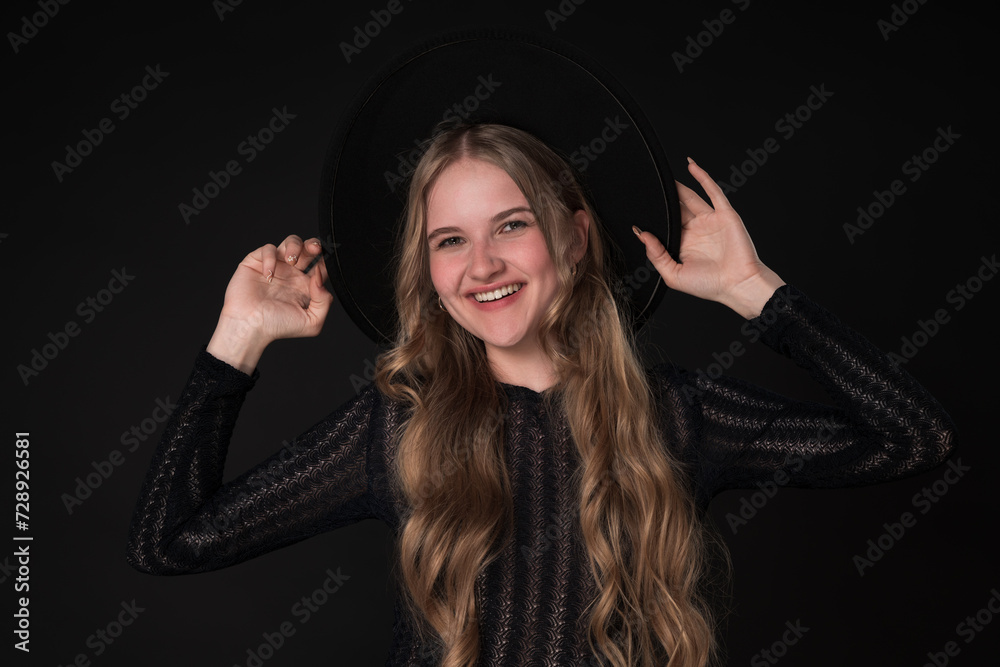 Portrait happy young woman with toothy smile looking at camera and holding brim of felt hat with both hands. Woman poses playfully on black background. Model with long hair dressed in black knit dress Portrait happy young woman with toothy smile looking at camera and holding brim of felt hat with both hands. Woman poses playfully on black background. Model with long hair dressed in black knit dress