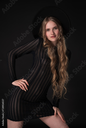 Blonde model exudes sensuality as she poses on black background, looking at camera. Woman wears chic, wide-brimmed felt hat, complete with black knit tight see-through dress that shows off her figure