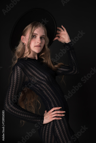 Stunning portrait features slim blonde fashion model exuding sensuality in relaxed pose on black background. Woman holds brim of hat with one hand and puts other hand on waist  dressed in black dress