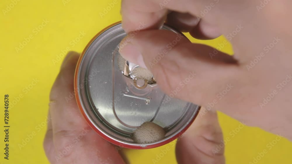 Above close-up shot: a hand opening a tin can, unleashing a burst of effervescent gas from a rich dark liquid carbonated drink, then taking the container away.