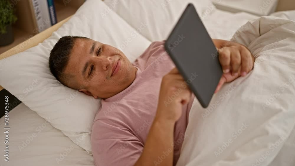 Happy young latin man enjoy relaxing morning on bed in bedroom, confident smiling while using touchpad, indoor portrait Happy young latin man enjoy relaxing morning on bed in bedroom, confident smiling while using touchpad, indoor portrait