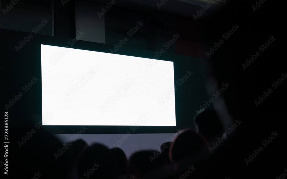 Mockup of big blank white screen with space in huge hall with scene and silhouette of audiences