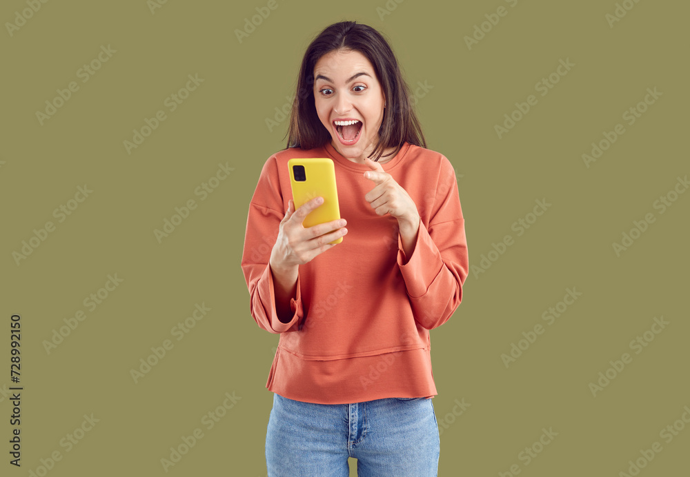 Wondered amazed excited happy young brunette woman in red sweatshirt and jeans looking at smartphone screen on khaki background. She is laughing with opened mouth and wide eyes. Amazing news concept. Wondered amazed excited happy young brunette woman in red sweatshirt and jeans looking at smartphone screen on khaki background. She is laughing with opened mouth and wide eyes. Amazing news concept.