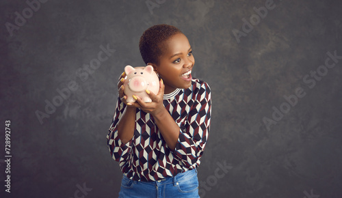 Studio portrait of happy smiling black woman holding pink piggy bank with savings to her ear, listening to sound of coins clinking inside, anticipating spending whole lot of money on fulfilling dreams photo