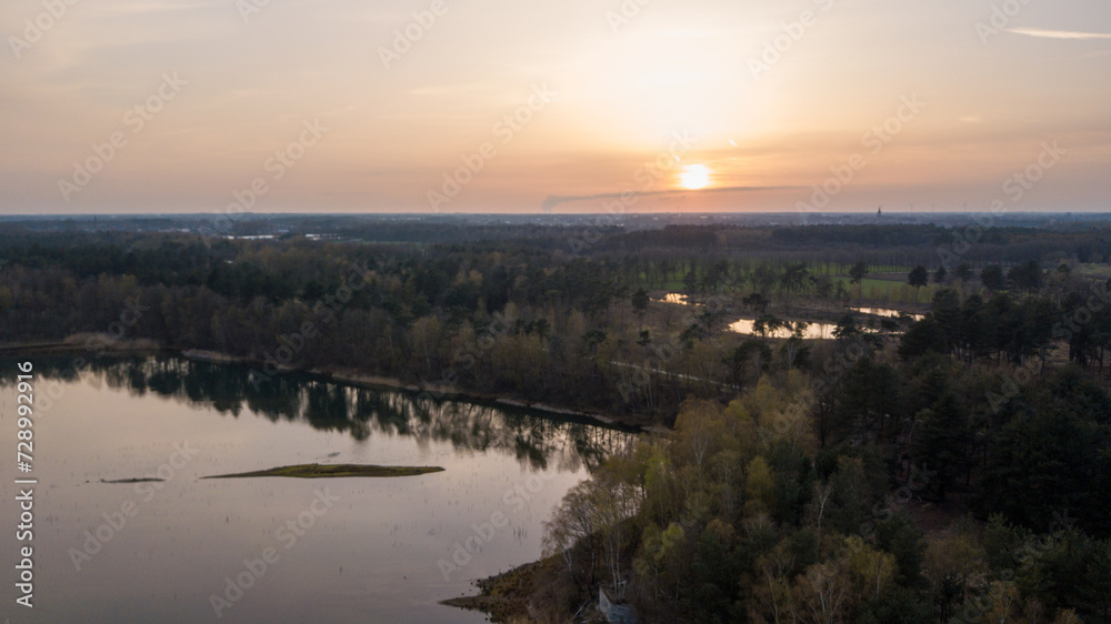 An aerial view captures the serene beauty of a forest lake at twilight. The setting sun dips towards the horizon, casting a gentle golden glow across the water's surface and the surrounding trees. The