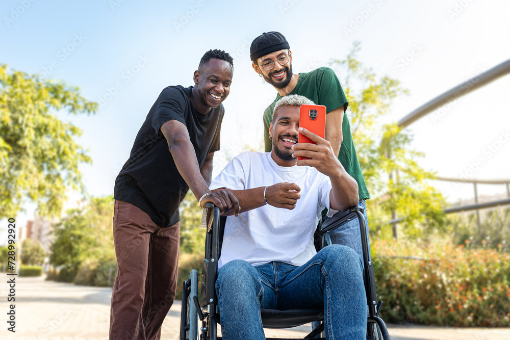 Fototapeta premium Young African American man in a wheelchair using phone together with male friends outdoors.