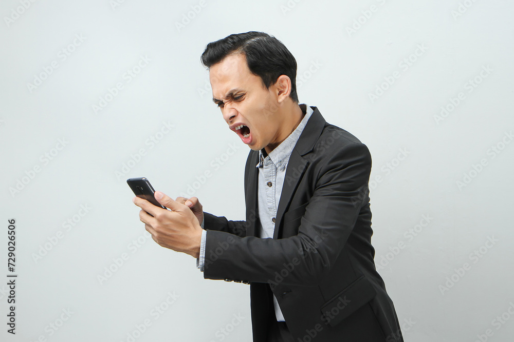 angry mad asian indonesian business man in suit holding smart phone on isolated background angry mad asian indonesian business man in suit holding smart phone on isolated background