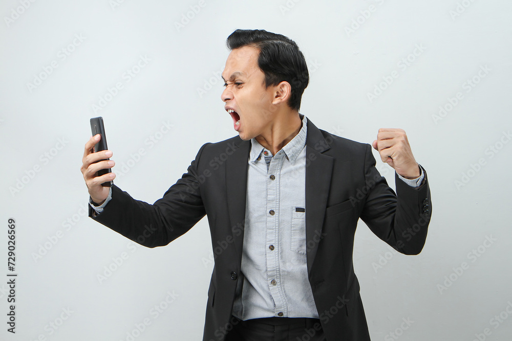 angry mad asian indonesian business man in suit holding smart phone on isolated background angry mad asian indonesian business man in suit holding smart phone on isolated background