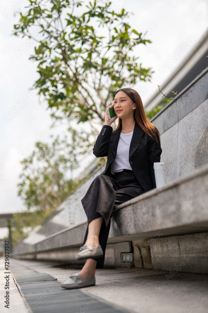 Fototapeta premium Asian businesswoman sitting and using phone outdoor, with a tumbler beside her
